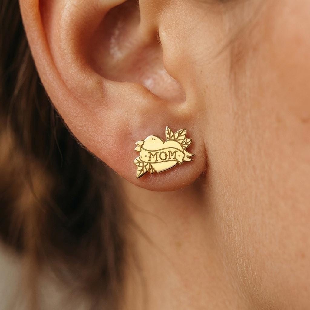 Pair of gold stud earrings shaped like a tattoo heart with a MOM banner