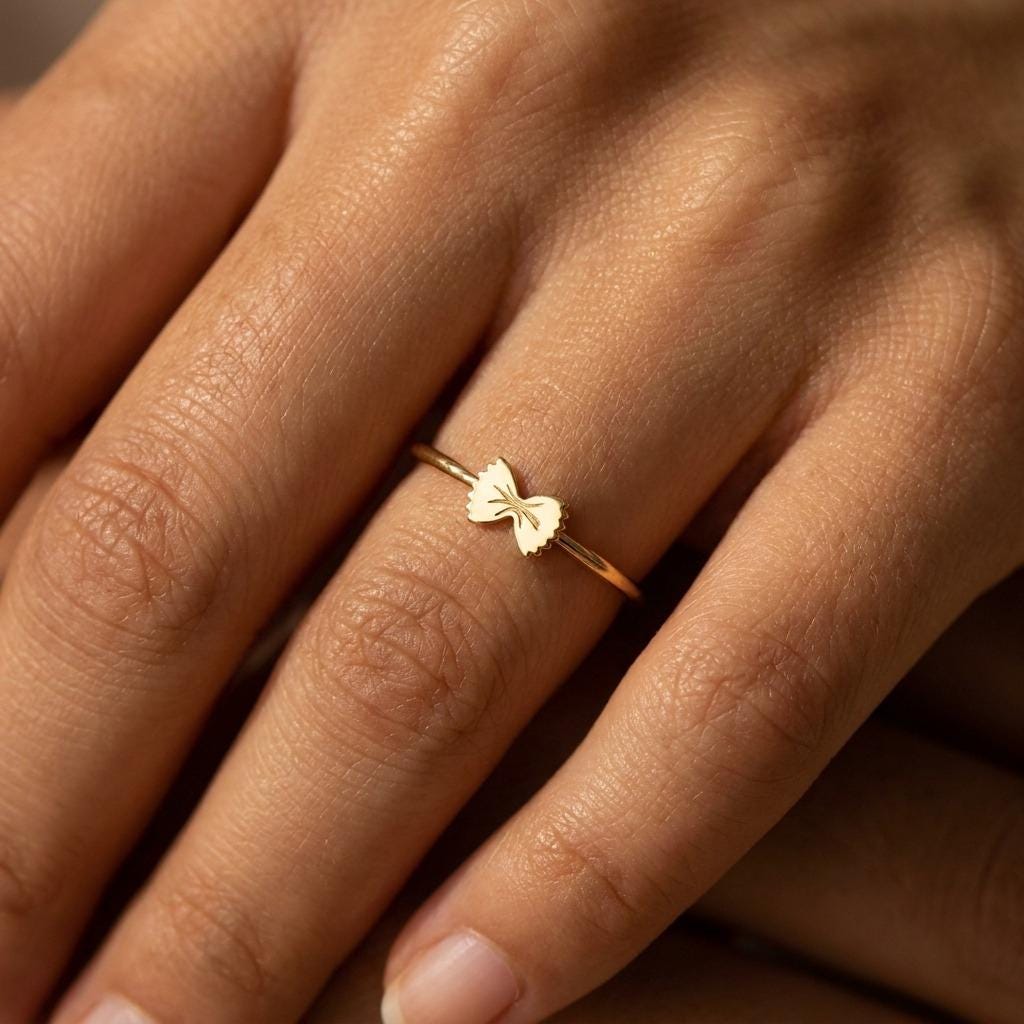 Thin gold ring topped with a small bowtie pasta shape, shown from above.
