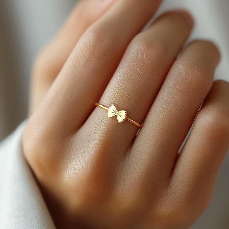 Thin gold ring topped with a small bowtie pasta shape, shown from above. (2 of 8)