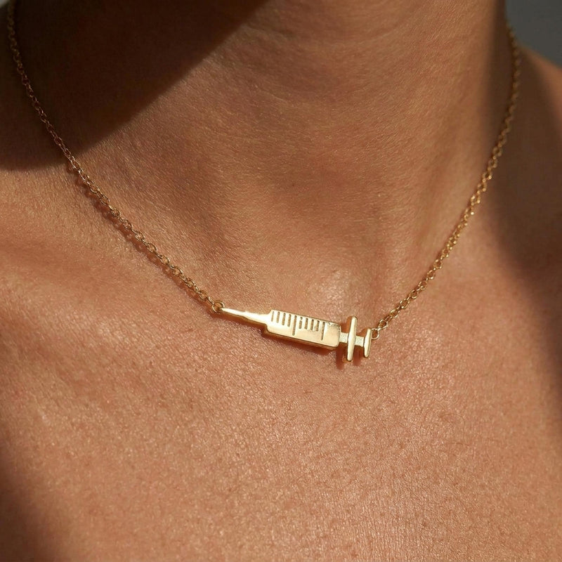 Slim gold syringe pendant with plunger and needle hanging from a thin chain
