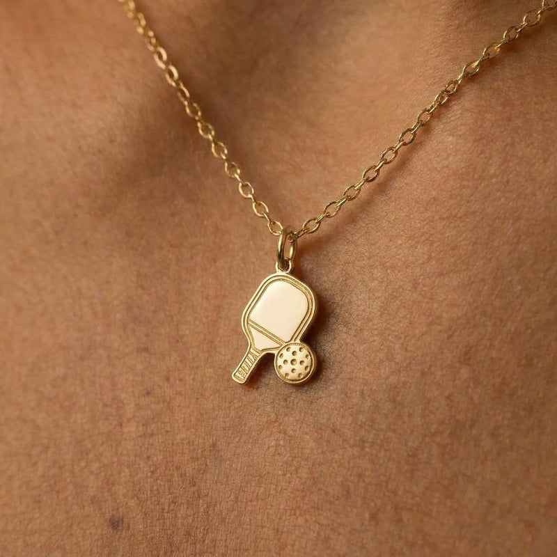 Small gold pickleball paddle pendant with perforated ball beside it on a chain