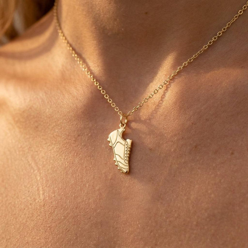 Small gold soccer cleat pendant hanging from a fine gold chain on a white background